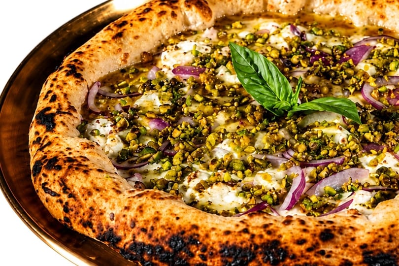 Close-up of a pizza with a golden crust, topped with pistachios, red onions, cheese, and basil.