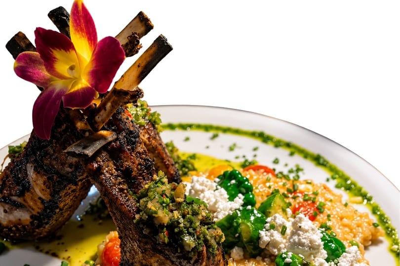 Grilled lamb chops with chimichurri, risotto, feta, and vegetables, garnished with an orchid.