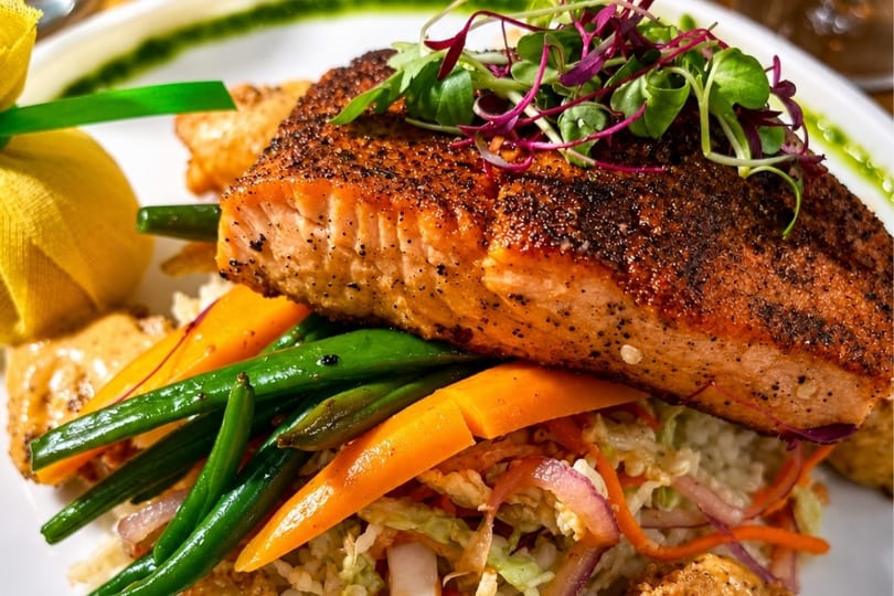Blackened salmon fillet with green beans, carrots, and coleslaw, garnished with microgreens.