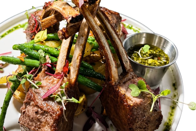 Grilled lamb chops with green beans, potatoes, and a side of pesto sauce on a white plate.